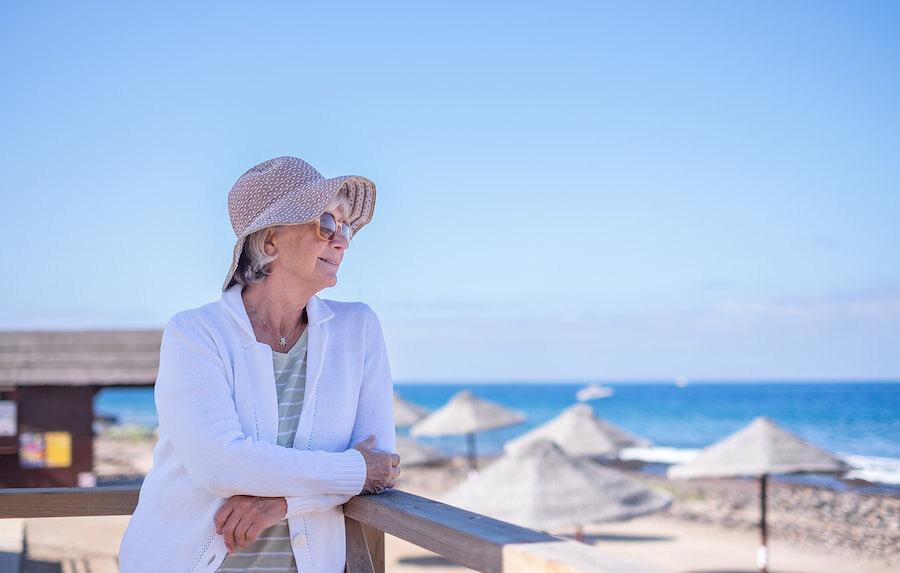 Femme âgée portant un chapeau de soleil et des lunettes de soleil, appuyée sur une rambarde en bois et regardant la mer. À l’arrière-plan, une plage ensoleillée avec des parasols en paille, un ciel bleu et des vagues calmes, évoquant une atmosphère paisible et détendue.