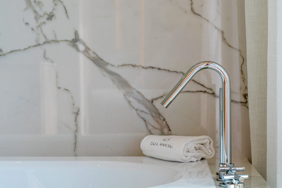 Close-up of a modern chrome bath tap set against a polished marble wall, with a folded Casa Marisal towel beside the bathtub.