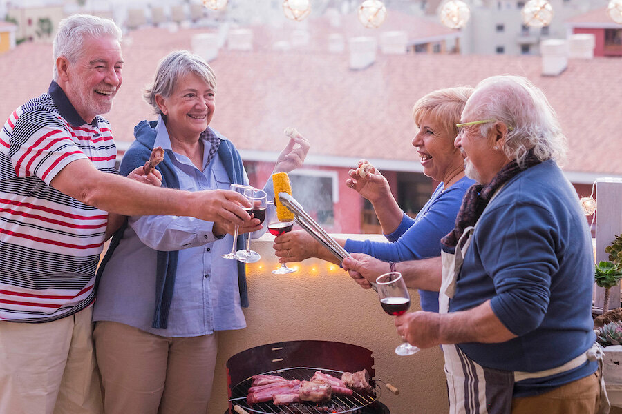 Groupe de quatre personnes âgées profitant d’un barbecue sur une terrasse. Elles sourient, tiennent des verres de vin rouge et cuisinent sur le gril. L’ambiance est chaleureuse et détendue, avec des guirlandes lumineuses et des toits visibles en arrière-plan.