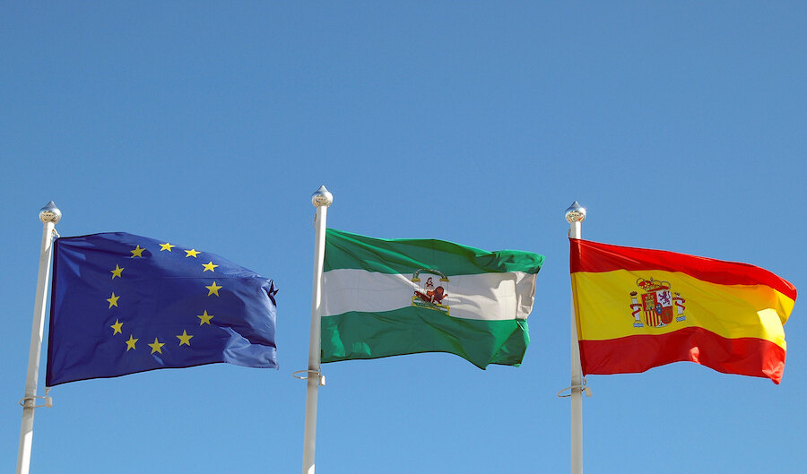 Flags of the European Union, Andalusia, and Spain waving against a clear blue sky.