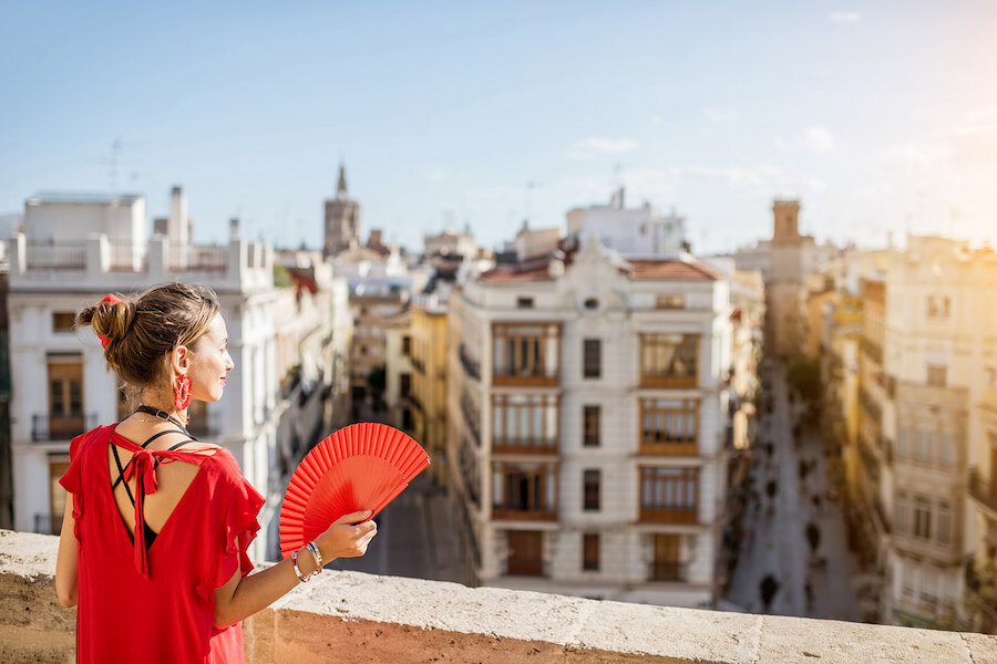 Woman dressed in red holding a red fan while overlooking a sunlit street and historic buildings in a Spanish city.