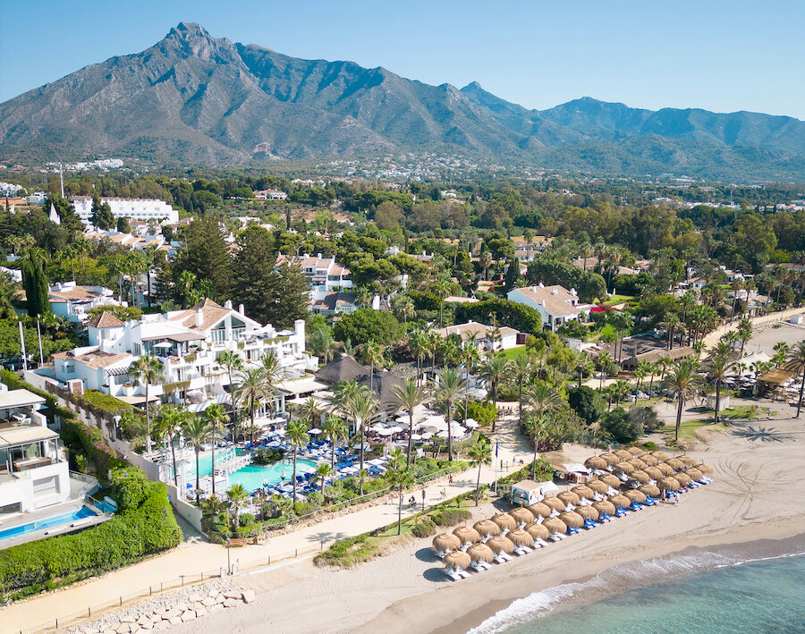 casa marisal. Aerial view of Marbella’s Golden Mile with the beach, thatched sunbeds, lush palm gardens, luxury beachfront resorts, and La Concha mountain rising in the background.