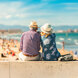 Couple de personnes âgées assis sur une promenade en bord de mer, portant des chapeaux d’été et regardant la plage. À l’arrière-plan, une côte ensoleillée animée de personnes profitant de la journée, avec la mer et des montagnes lointaines sous un ciel bleu éclatant. L’image évoque la détente, la complicité et le style de vie méditerranéen.
