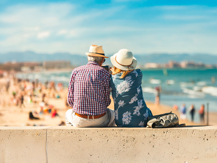 Ouder koppel dat op een promenade aan zee zit, met zomerhoeden op en uitkijkend over het strand. Op de achtergrond is een zonnige kustlijn te zien met mensen die van de dag genieten, de zee en verre bergen onder een helderblauwe lucht. De afbeelding straalt ontspanning, gezelschap en de mediterrane levensstijl uit.