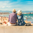 Elderly couple sitting on a seaside promenade, wearing summer hats and looking out at the beach. The background shows a sunny coastline filled with people enjoying the day, with the sea and distant mountains under a bright blue sky. The image conveys relaxation, companionship, and the Mediterranean lifestyle.