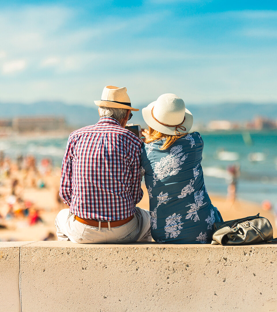 Couple de personnes âgées assis sur une promenade en bord de mer, portant des chapeaux d’été et regardant la plage. À l’arrière-plan, une côte ensoleillée animée de personnes profitant de la journée, avec la mer et des montagnes lointaines sous un ciel bleu éclatant. L’image évoque la détente, la complicité et le style de vie méditerranéen.