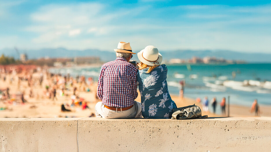 Pareja de personas mayores sentada en un paseo marítimo, con sombreros de verano y mirando hacia la playa. Al fondo se aprecia una costa soleada con gente disfrutando del día, el mar y montañas a lo lejos bajo un cielo azul brillante. La imagen transmite relajación, compañía y el estilo de vida mediterráneo.
