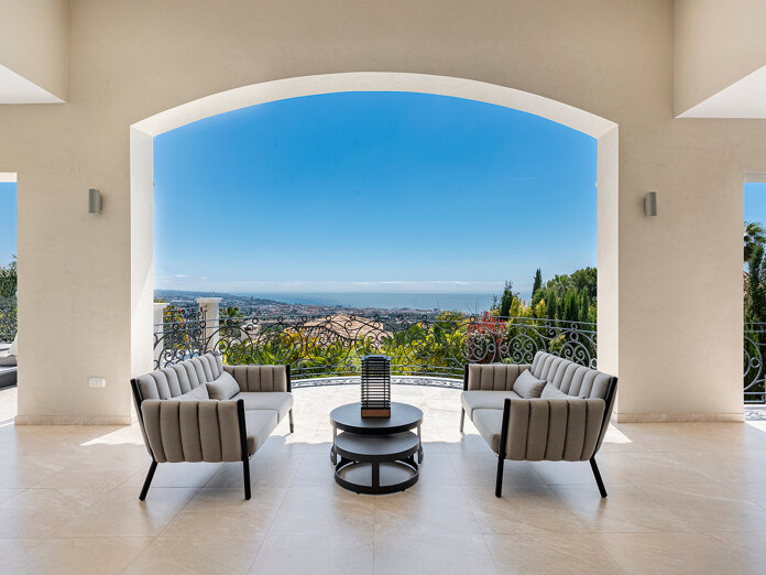 Überdachte Terrassenlounge mit modernen Sesseln, rundem Tisch und schmiedeeisernem Geländer, mit Blick auf Marbella, üppige Gärten und den Panoramablick auf das Mittelmeer unter einem wolkenlosen blauen Himmel.