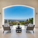 Covered terrace lounge with modern armchairs, round table and wrought-iron balustrade overlooking Marbella, lush gardens and panoramic Mediterranean Sea views under a clear blue sky.