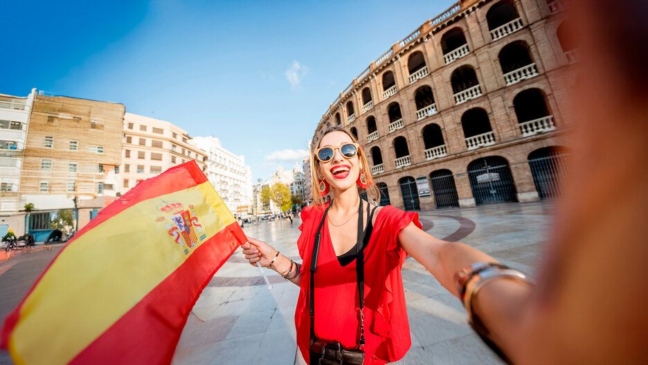 Femme souriante vêtue de rouge tenant un drapeau espagnol et prenant un selfie devant un bâtiment historique par une journée ensoleillée en Espagne.