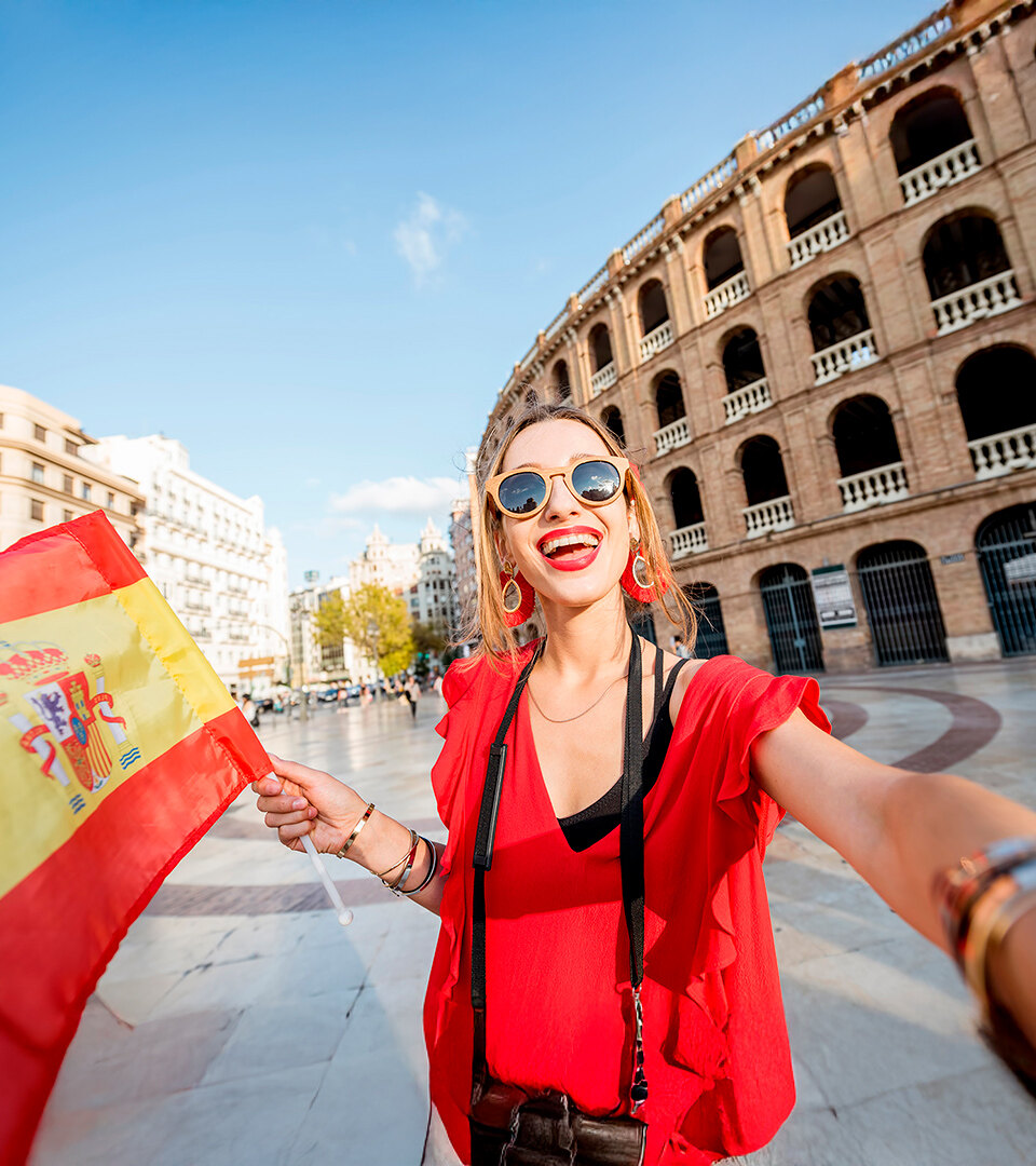 Smiling woman wearing red clothes holding a Spanish flag and taking a selfie in front of a historic building on a sunny day in Spain.