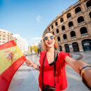 Smiling woman wearing red clothes holding a Spanish flag and taking a selfie in front of a historic building on a sunny day in Spain.