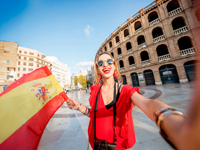 Femme souriante vêtue de rouge tenant un drapeau espagnol et prenant un selfie devant un bâtiment historique par une journée ensoleillée en Espagne.