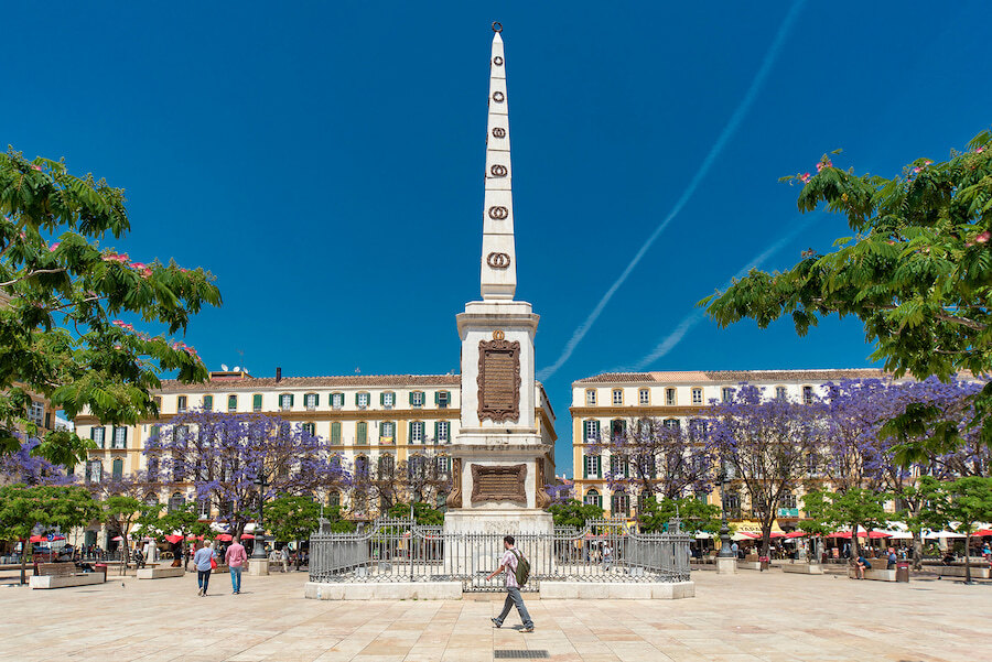 Plaza de la Merced in Málaga, featuring the central obelisk monument surrounded by jacaranda trees in bloom, historic buildings with colourful facades, and people enjoying the lively atmosphere under a bright blue sky.