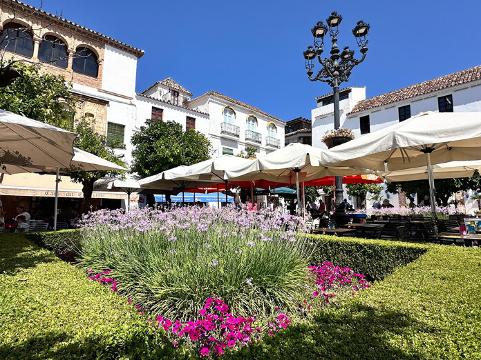 Sunny day at Plaza de los Naranjos in Marbella’s Old Town, featuring outdoor café umbrellas, blooming lavender and pink flowers, traditional white Andalusian buildings, and a wrought-iron street lamp against a bright blue sky.