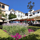 Sunny day at Plaza de los Naranjos in Marbella’s Old Town, featuring outdoor café umbrellas, blooming lavender and pink flowers, traditional white Andalusian buildings, and a wrought-iron street lamp against a bright blue sky.