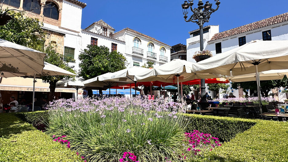 Sunny day at Plaza de los Naranjos in Marbella’s Old Town, featuring outdoor café umbrellas, blooming lavender and pink flowers, traditional white Andalusian buildings, and a wrought-iron street lamp against a bright blue sky.