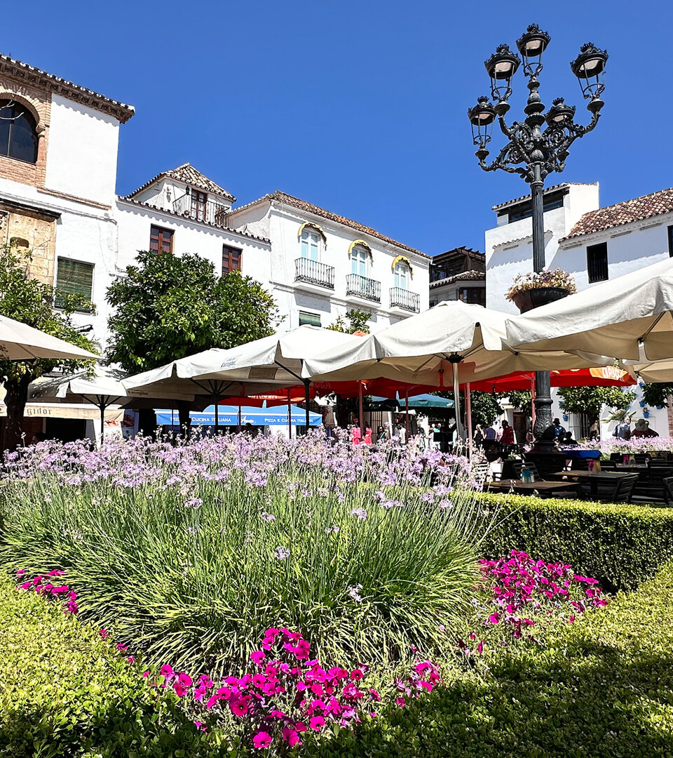 Sunny day at Plaza de los Naranjos in Marbella’s Old Town, featuring outdoor café umbrellas, blooming lavender and pink flowers, traditional white Andalusian buildings, and a wrought-iron street lamp against a bright blue sky.