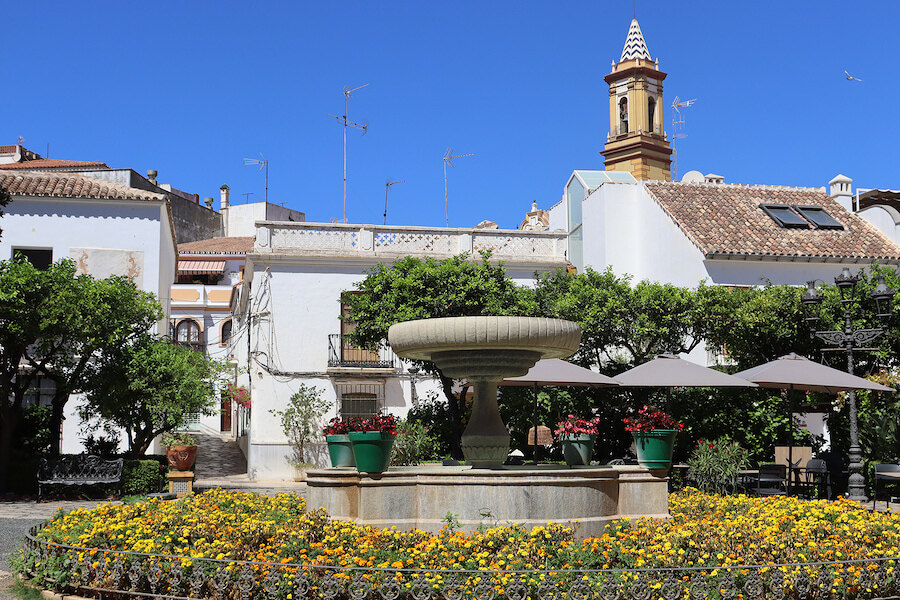 Picturesque view of Plaza de las Flores in Estepona, featuring a central stone fountain surrounded by vibrant yellow flowers, lush greenery, and traditional Andalusian whitewashed buildings under a clear blue sky. prettiest plazas