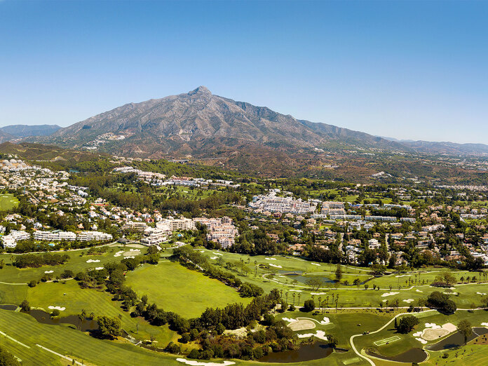 Vue aérienne panoramique de Nueva Andalucía à Marbella, mettant en valeur ses terrains de golf luxuriants, ses lotissements résidentiels et les collines verdoyantes environnantes, avec le sommet saisissant de la montagne La Concha dominant en arrière-plan sous un ciel bleu clair.
