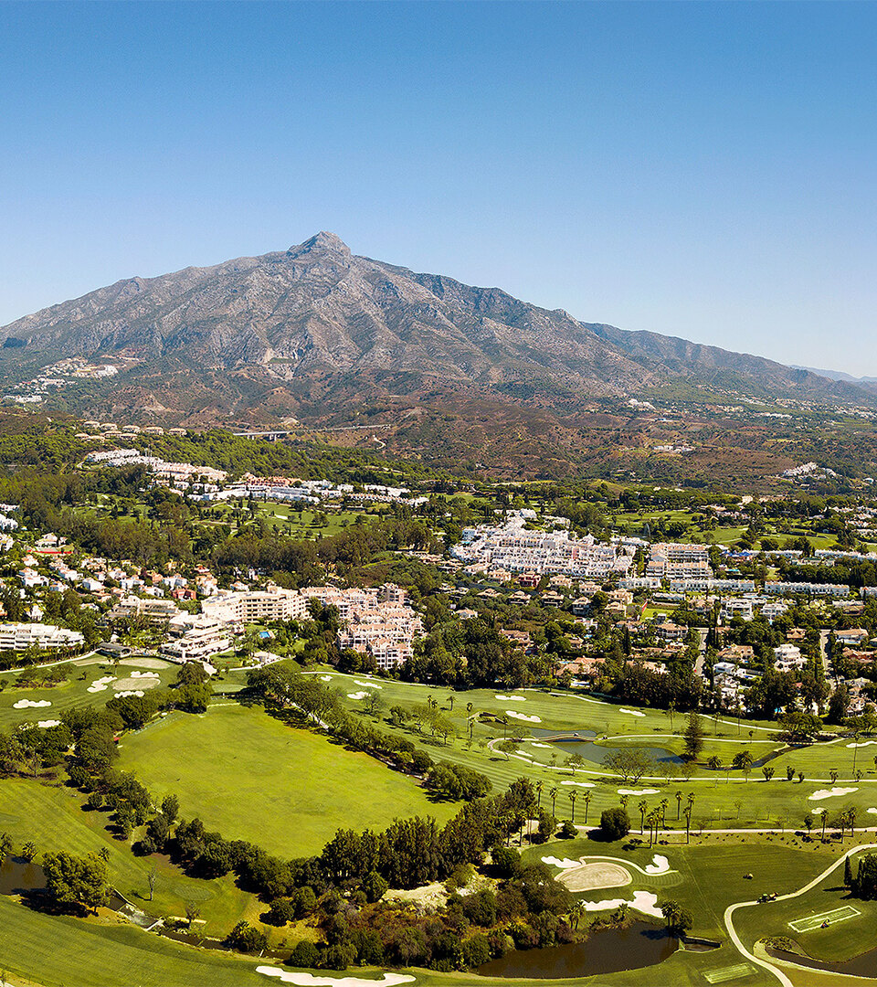 Vista aérea panorámica de Nueva Andalucía en Marbella, que muestra sus exuberantes campos de golf, desarrollos residenciales y las verdes colinas circundantes, con el impresionante pico de la montaña La Concha elevándose al fondo bajo un cielo azul claro.