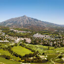 Panoramische Luftaufnahme von Nueva Andalucía in Marbella mit üppigen Golfplätzen, Wohnanlagen und den umliegenden grünen Hügeln, im Hintergrund der markante Gipfel des La-Concha-Berges unter einem klaren blauen Himmel.