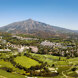 Vista aérea panorámica de Nueva Andalucía en Marbella, que muestra sus exuberantes campos de golf, desarrollos residenciales y las verdes colinas circundantes, con el impresionante pico de la montaña La Concha elevándose al fondo bajo un cielo azul claro.