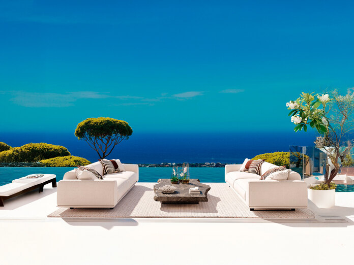 Luxe buitenterras met witte banken, een infinity pool en een panoramisch uitzicht op de Middellandse Zee en weelderig groen onder een helderblauwe hemel.