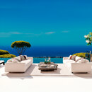 Luxuriöse Außenterrasse mit weißen Sofas, einem Infinity-Pool und Panoramablick auf das Mittelmeer und üppiges Grün unter einem klaren blauen Himmel.