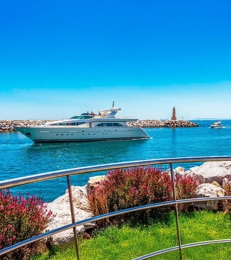Een luxe jacht vaart door het helderblauwe water van een jachthaven in Marbella, met een rotsachtige golfbreker en een vuurtoren op de achtergrond. Op de voorgrond zie je verzorgd groen gras, felroze bloemen en een strakke roestvrijstalen reling onder een strakblauwe lucht.