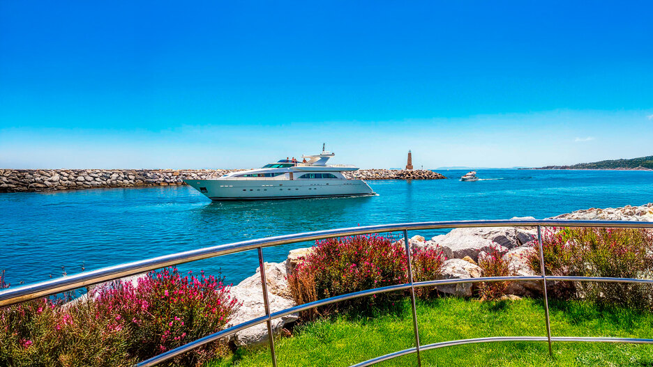 A luxury yacht sails through the bright blue waters of a marina in Marbella, with a rocky breakwater and lighthouse in the background. The foreground features manicured green grass, vibrant pink flowers, and a sleek stainless-steel railing under a clear blue sky.