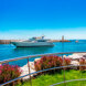 Un yacht de luxe vogue sur les eaux bleu vif d'une marina de Marbella, avec une digue rocheuse et un phare en arrière-plan. Au premier plan, une pelouse verte impeccable, des fleurs rose vif et une élégante rambarde en acier inoxydable sous un ciel bleu azur.