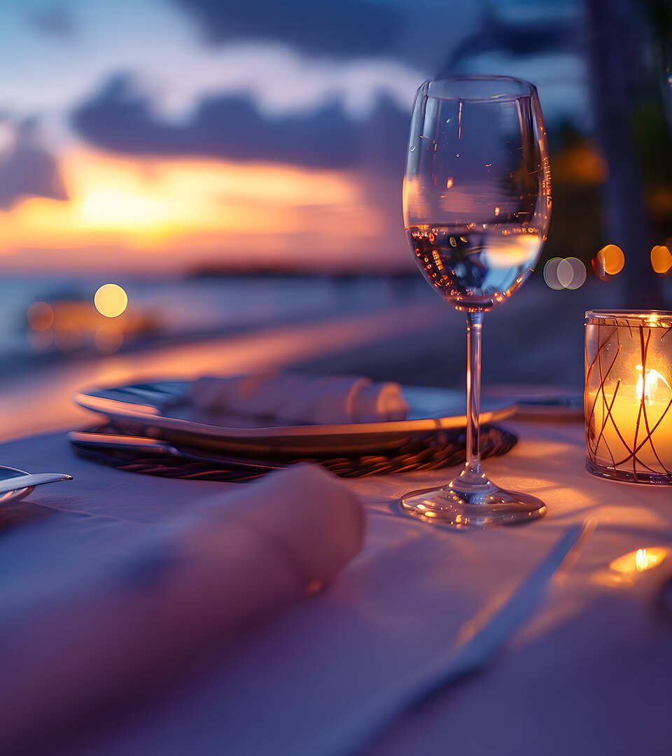 Romantic seaside dining table set for two at sunset, featuring a wine glass, lit candle, and elegant tableware with a blurred view of the ocean and colorful sky in the background.