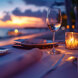 Romantic seaside dining table set for two at sunset, featuring a wine glass, lit candle, and elegant tableware with a blurred view of the ocean and colorful sky in the background.
