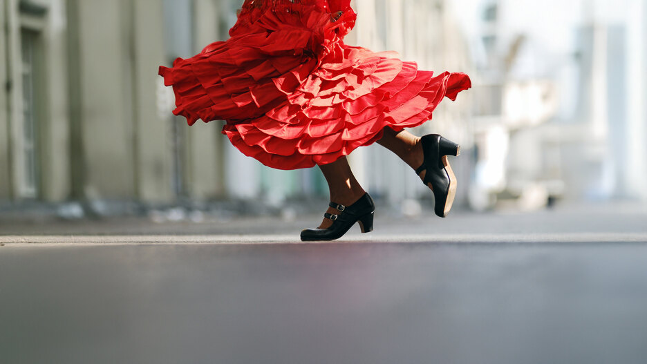 Primer plano de una bailarina de flamenco en movimiento, con un vibrante vestido rojo con volantes que gira y zapatos de tacón negros tradicionales a medio paso, capturando la energía y la elegancia de la danza española.