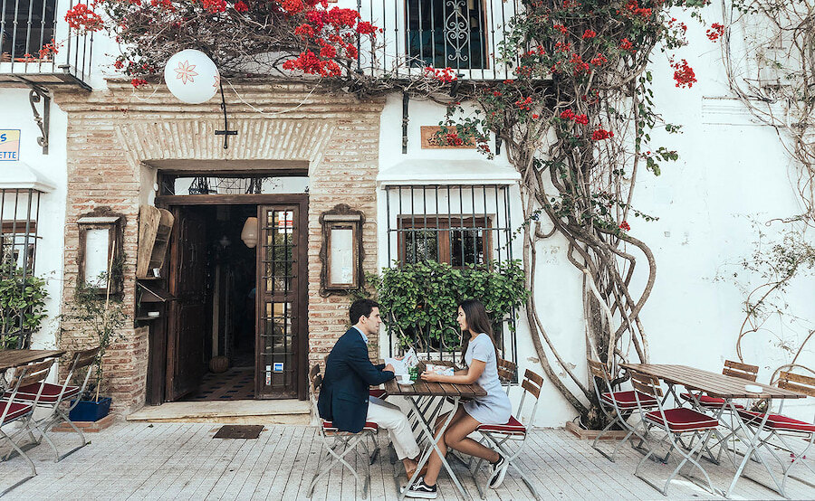A couple dining outdoors at Casanis Bistrot restaurant, seated at a small wooden table on a cobblestone street, with white walls and red flowers climbing up the building facade in the background.