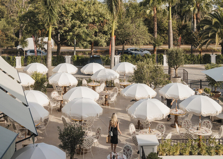 Outdoor terrace of Florentine restaurant at The Forum Marbella, featuring white parasols over round tables with decorative chairs, surrounded by greenery and palm trees, with a woman in a black dress walking through the area