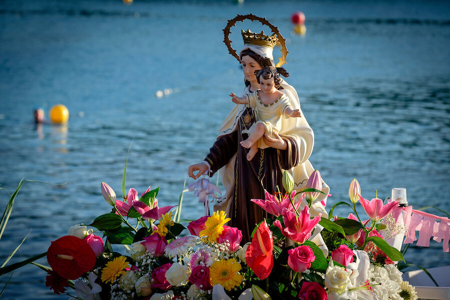 Statue der Jungfrau Maria mit dem Jesuskind im Arm, geschmückt mit einer goldenen Krone und umgeben von einem farbenfrohen Arrangement aus frischen Blumen, vor der Kulisse eines ruhigen blauen Meeres während einer traditionellen maritimen religiösen Feier.
