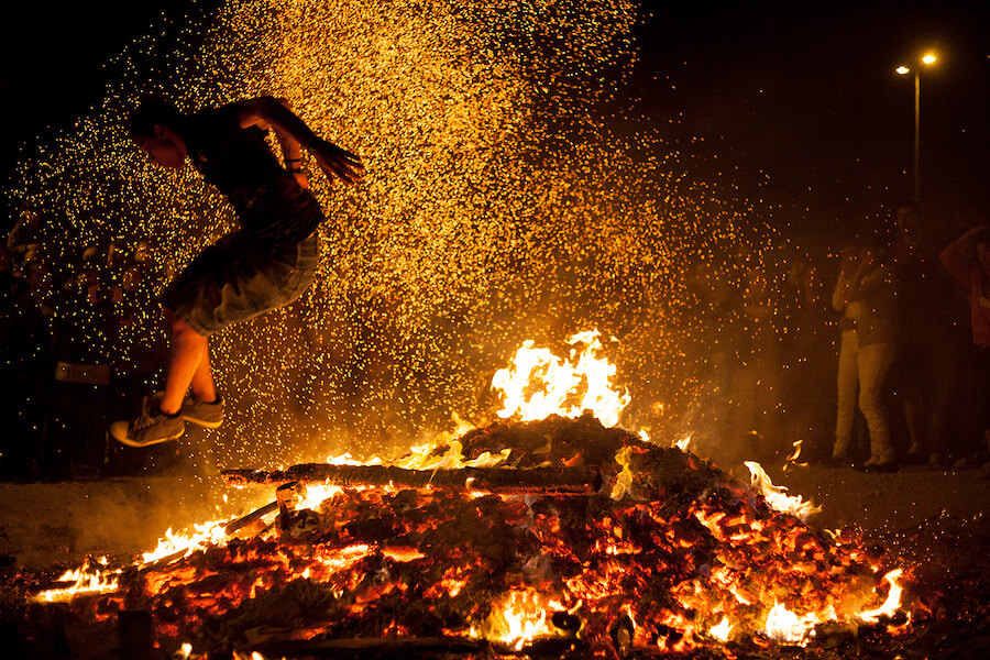 Ein junger Mann springt nachts mitten in der Luft über ein loderndes Lagerfeuer, während die Funken dramatisch in alle Richtungen fliegen. Im Hintergrund beobachtet eine Menschenmenge, die sich vor dem Schein der Flammen abhebt und die Intensität und Tradition eines Feuersprung-Festes oder einer Feier weckt.