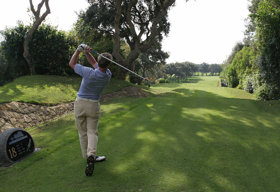 Een golfer rondt zijn swing af op de 18e tee van een weelderige, met bomen omzoomde golfbaan, omringd door groen en gevlekt zonlicht op een rustige dag.