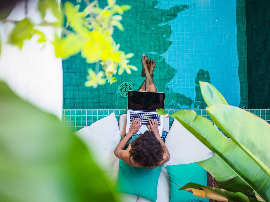 Bovenaanzicht van een persoon die op een laptop werkt, terwijl hij ontspant op een ligstoel met kussens, met zijn benen in een zwembad met turquoise tegels, omringd door tropische planten en natuurlijk zonlicht. Dit is de belichaming van de levensstijl van de digitale nomaden.