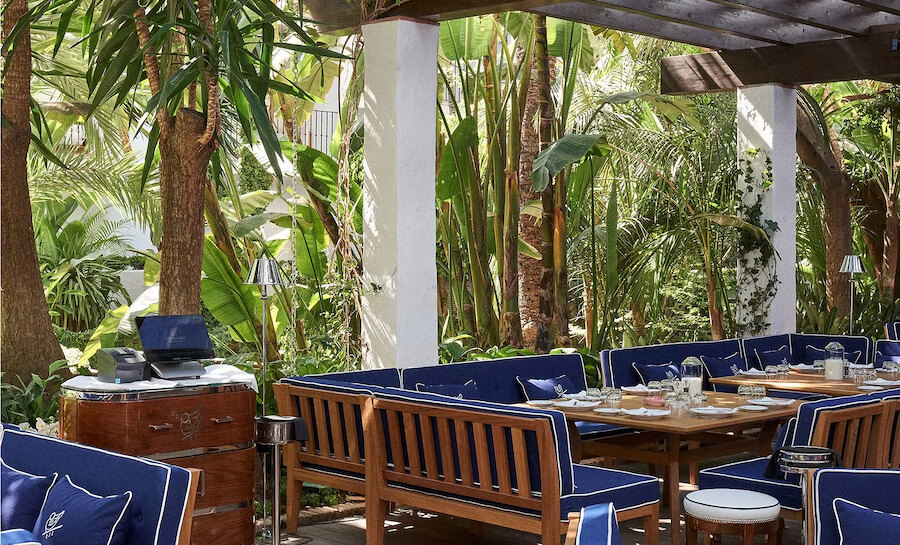 Outdoor dining area at Cipriani restaurant in Puente Romano hotel, featuring wooden tables and chairs with blue cushions, surrounded by lush tropical plants and shaded by a pergola.