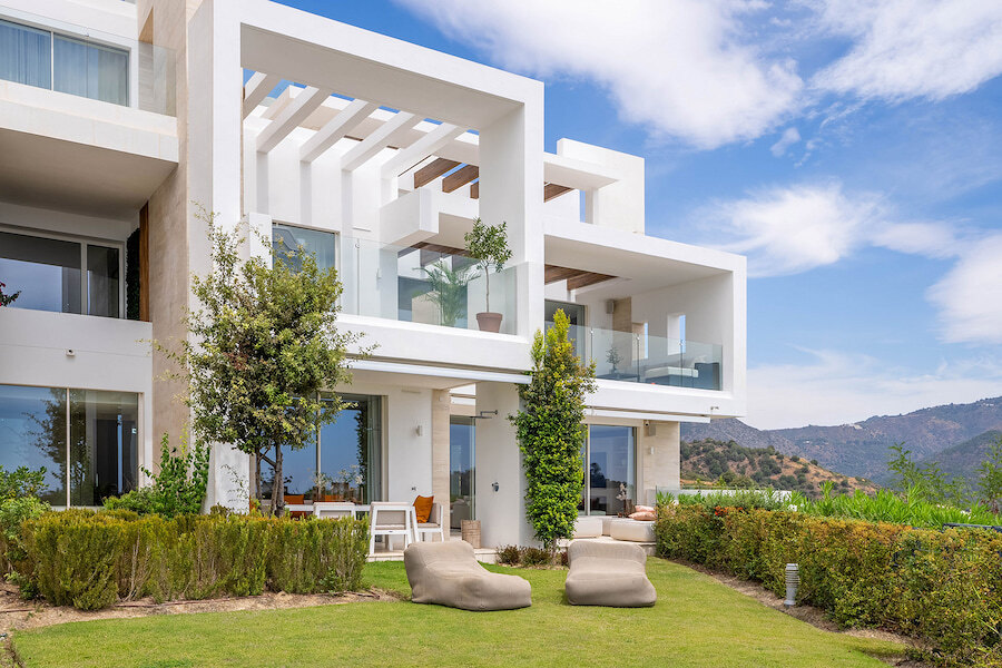 Modernes Apartmentgebäude in Palo Alto in Ojén, Marbella, mit klarer weißer Architektur, geräumigen Terrassen mit Glasbalustraden, angelegten Gärten und malerischer Aussicht auf die umliegenden Hügel und Berge.