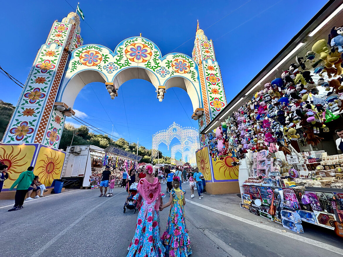 Farbenfroher Eingangsbogen zu einem traditionellen andalusischen Jahrmarkt, dekoriert mit Blumenmotiven und hellen Lichtern. Menschen schlendern über das von Ständen gesäumte Festgelände, an dem Spielzeug, Süßigkeiten und Souvenirs verkauft werden. Zwei Frauen in leuchtenden Flamencokleidern gehen Hand in Hand und tragen zur festlichen Atmosphäre unter einem strahlend blauen Himmel bei.