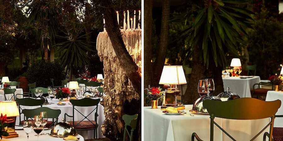 Evening outdoor dining at the Grill restaurant of Marbella Club, featuring elegantly set tables with white tablecloths, wine glasses, lamps, and surrounded by lush greenery and trees.
