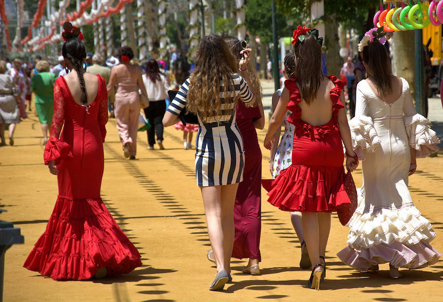 Vrouwen gekleed in traditionele flamencojurken en feestelijke kledij lopen over een versierd kermispad tijdens een Spaanse feria, met kleurrijke slingers boven hun hoofd en mensen die op de achtergrond genieten van het levendige culturele evenement.