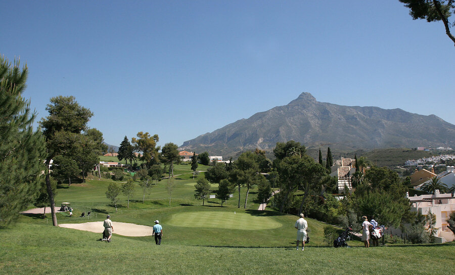 Golfistas en un exuberante campo verde en el Valle del Golf de Nueva Andalucía, Marbella, con calles cuidadas rodeadas de árboles y villas, y la impresionante montaña La Concha elevándose al fondo bajo un cielo azul claro.