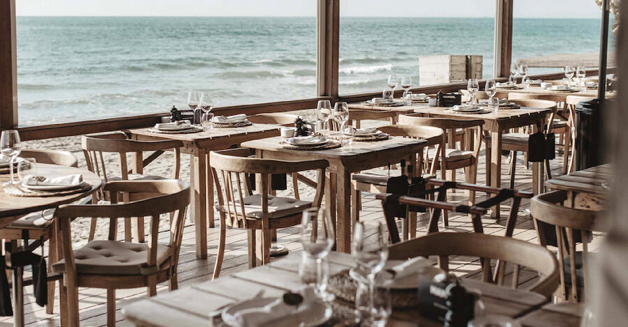 Indoor dining area at Le Casanis Beach Club in Marbella, featuring rustic wooden tables and chairs set with wine glasses and napkins, with large windows offering a view of the sea and sandy beach.