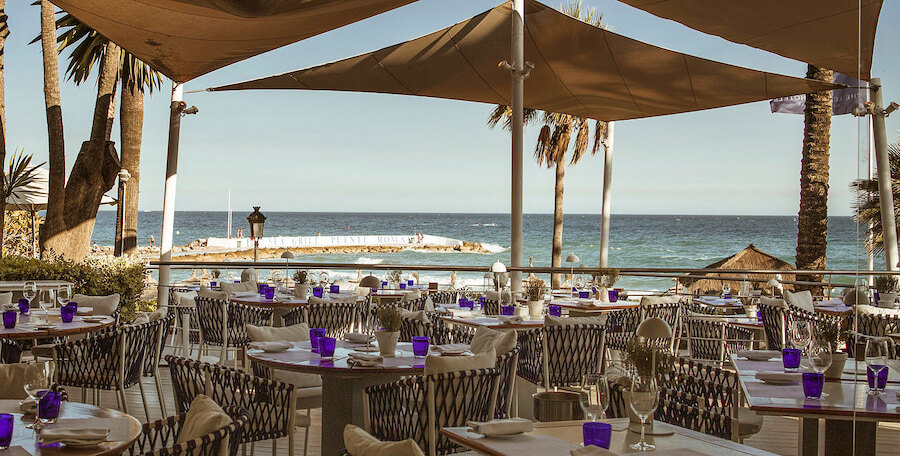 terrace dining area at Sea Grill restaurant in Puente Romano Beach Resort, with tables set with white tablecloths, purple glasses, and cutlery, overlooking the sea with palm trees and a sunny sky