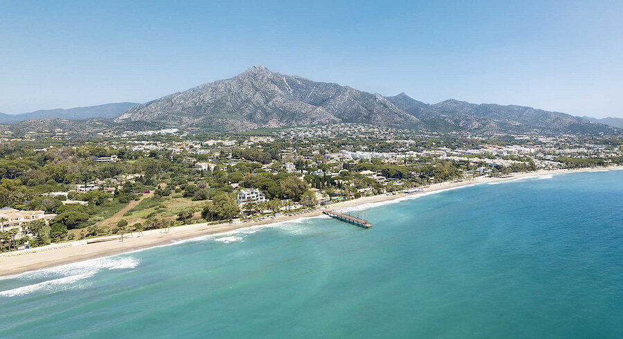 Luchtfoto van de Golden Mile van Marbella, met het turquoise water van de Middellandse Zee, een zandstrand met een pier en weelderig groen dat zich uitstrekt tot aan de uitlopers van de berg La Concha onder een helderblauwe hemel.